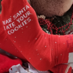 Person wearing red socks that read "Dear Santa, I ate your cookies" next to a plate of cookies.