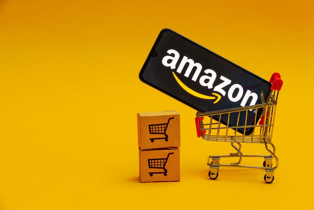 Amazon logo on a smartphone screen inside a toy shopping cart with a yellow background, representing the growth of e-commerce.