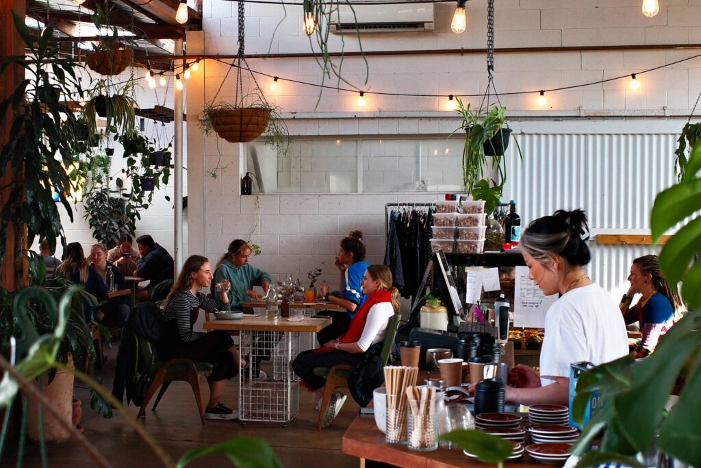 A busy, indoor Adelaide café with patrons sitting at tables, string lights overhead, and an employee working at the counter.