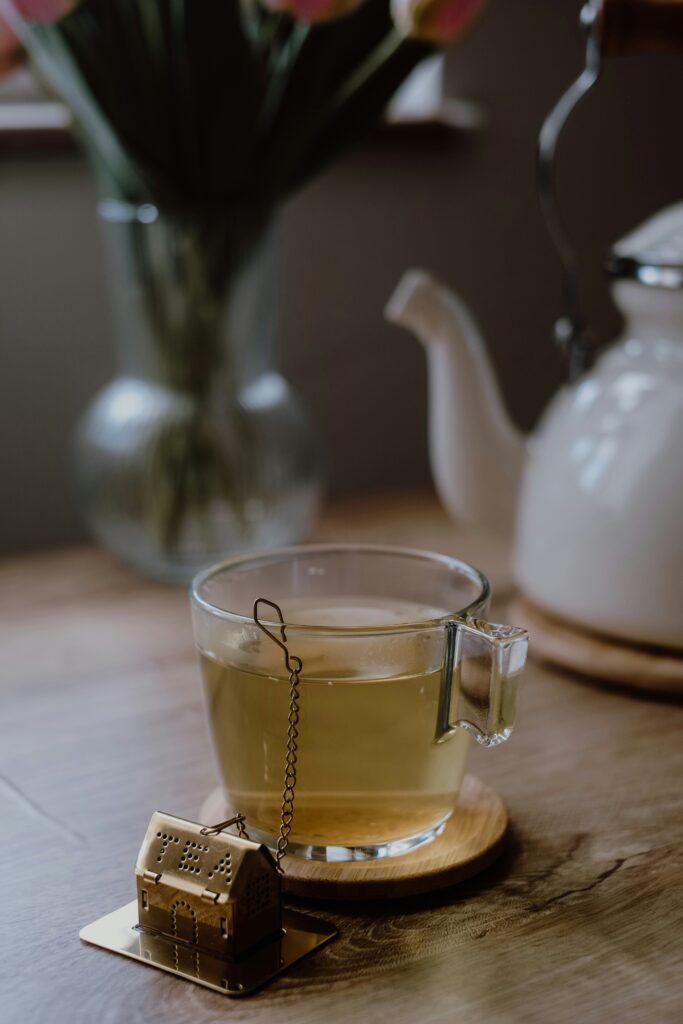 A clear glass mug of light-colored tea on a wooden coaster, next to a gold house-shaped tea infuser, with a white teapot in the background.