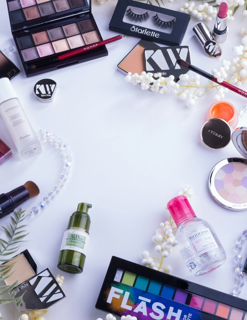 Flat lay of various cosmetic and skincare products, including eyeshadow palettes, false lashes, makeup remover, and lipstick, surrounding a central white space.