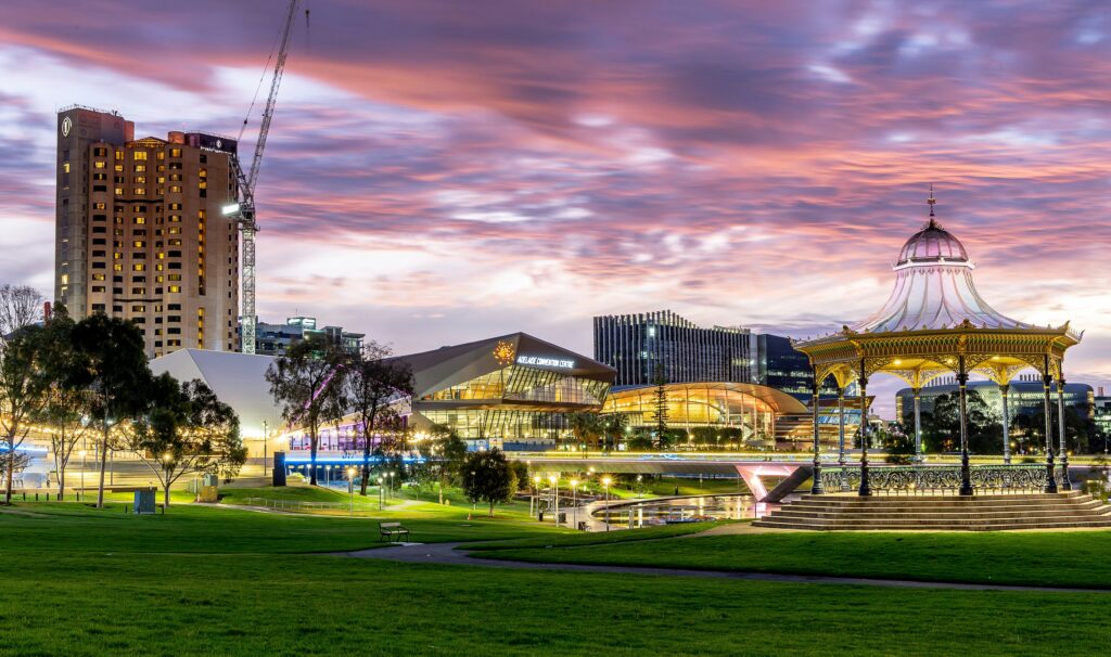 Panoramic view of Adelaide city at dusk, showing modern buildings, a lush green park, and a prominent gold rotunda under a pink and purple sky.