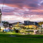 Panoramic view of Adelaide city at dusk, showing modern buildings, a lush green park, and a prominent gold rotunda under a pink and purple sky.