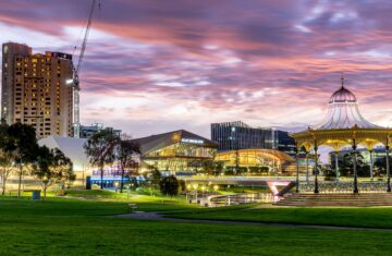 Panoramic view of Adelaide city at dusk, showing modern buildings, a lush green park, and a prominent gold rotunda under a pink and purple sky.
