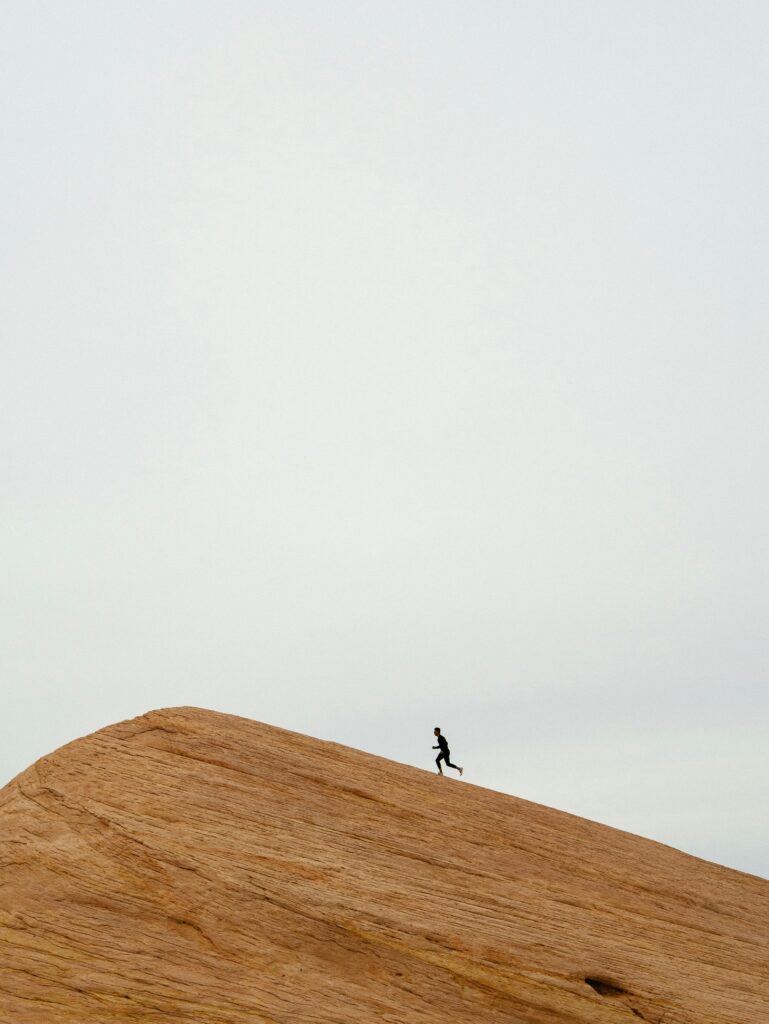 Silhouette of a lone runner ascending a large, bare, textured sand-colored hill against a bright sky.