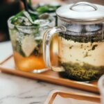 Close-up of a glass infuser mug with brewing loose leaf tea next to a tall glass of iced tea with lime and mint on a wooden tray.