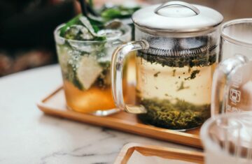Close-up of a glass infuser mug with brewing loose leaf tea next to a tall glass of iced tea with lime and mint on a wooden tray.