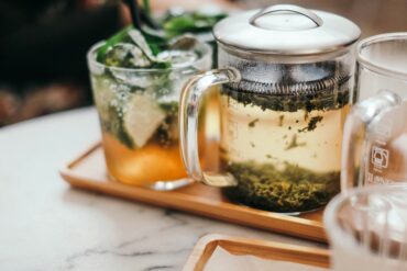 Close-up of a glass infuser mug with brewing loose leaf tea next to a tall glass of iced tea with lime and mint on a wooden tray.