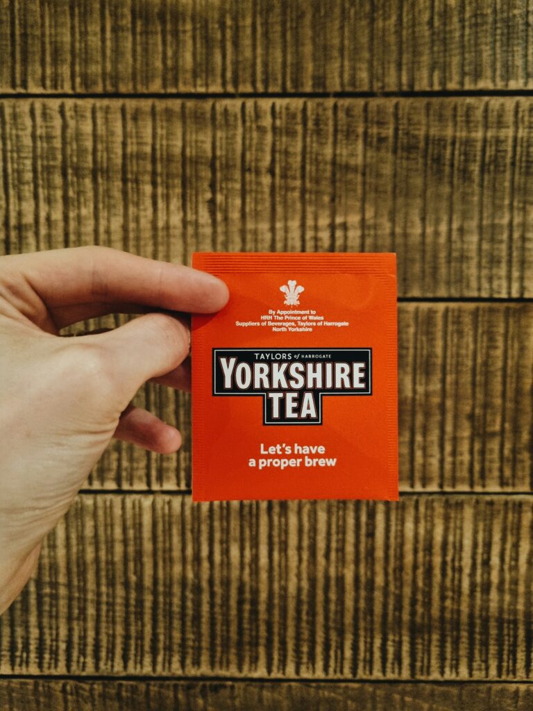 A hand holding a single orange packet of Yorkshire Tea against a wooden background.