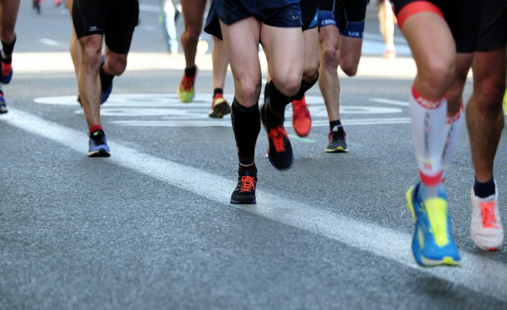 Close-up of runners' legs and feet in a marathon or race, showing various types of running shoes on the asphalt.