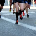 Close-up of runners' legs and feet in a marathon or race, showing various types of running shoes on the asphalt.