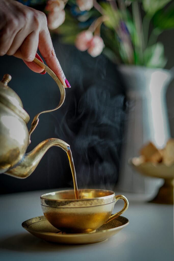 A hand pouring dark, steaming hot tea from a vintage brass teapot into a gold-plated teacup and saucer.
