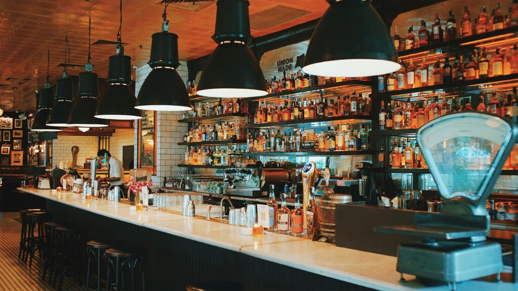 A long marble bar counter with black stools, brightly lit by hanging pendant lights, and shelves filled with hundreds of liquor bottles in the background.