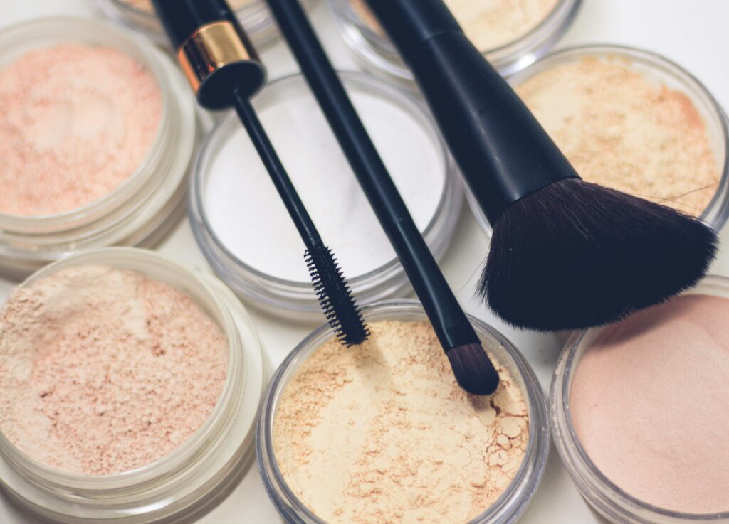 Close-up of several open jars of loose powder makeup in various skin tones, with a mascara wand and two makeup brushes laid on top.