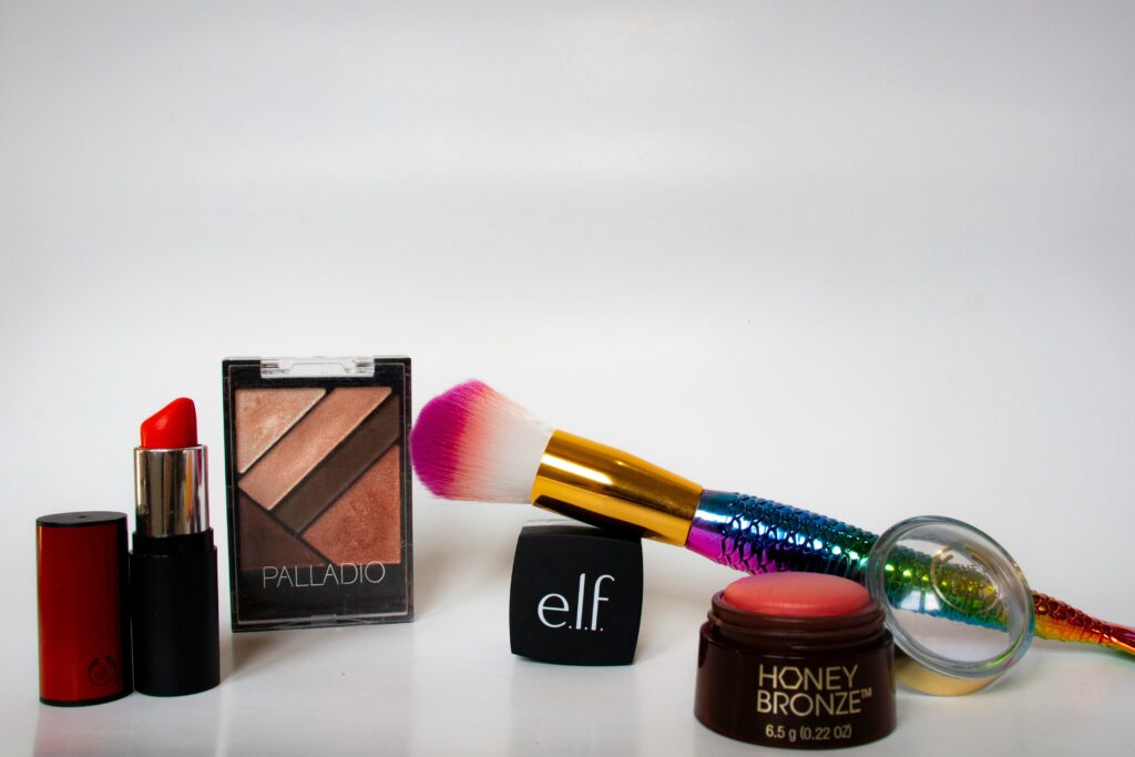 A bright, simple shot of various cosmetics, including a lipstick, eyeshadow palette, and a pink and white makeup brush, next to a black cube with the 'e.l.f.' logo.