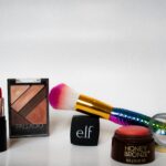 A bright, simple shot of various cosmetics, including a lipstick, eyeshadow palette, and a pink and white makeup brush, next to a black cube with the 'e.l.f.' logo.