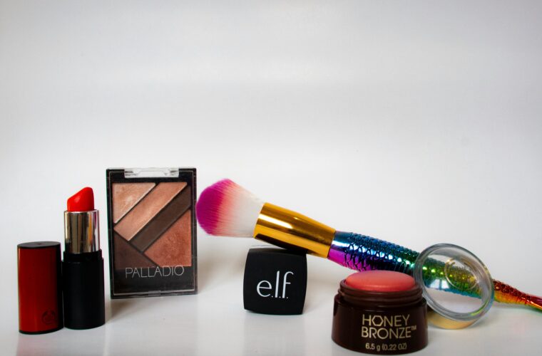 A bright, simple shot of various cosmetics, including a lipstick, eyeshadow palette, and a pink and white makeup brush, next to a black cube with the 'e.l.f.' logo.