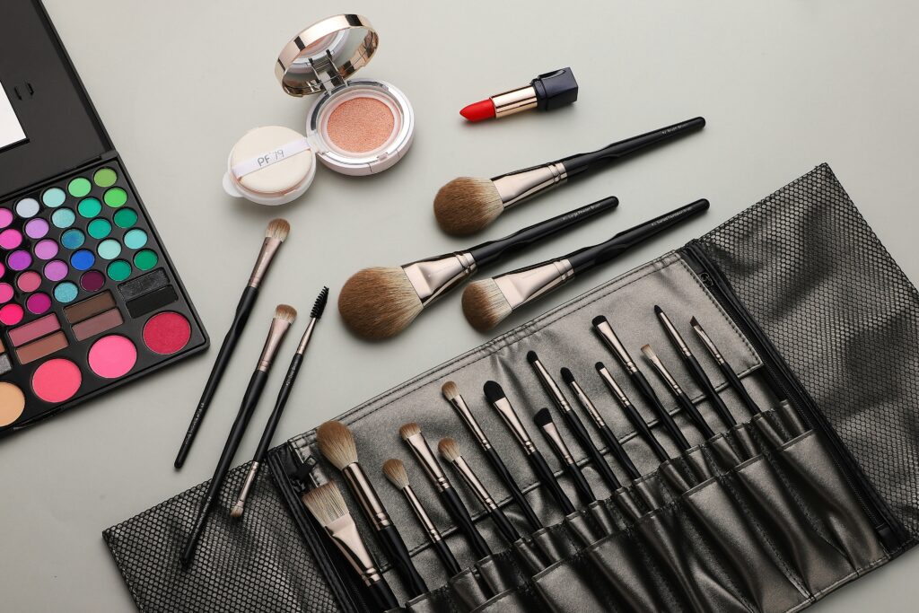 An array of professional makeup brushes, including a set organized in a black faux leather case, next to a colorful eyeshadow palette and a red lipstick.