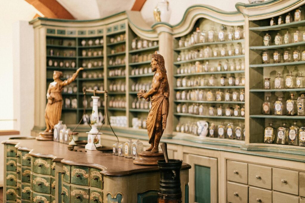 Interior of an old-fashioned apothecary with rows of glass jars on ornate green shelves and wooden statues on a counter.